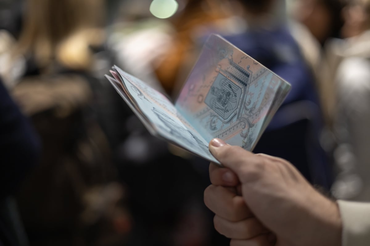 A close-up, low-angle shot of a person's hand holding a travel passport, open to the inner pages showing the national crest, possibly while queuing or going through border control at an airport